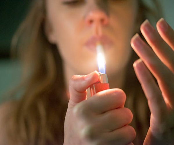 Grunge dark portrait of woman smoking. Girl lighting cigarette indoors at her living room, close up,   female fingers holding cigarette and lighter fire. concept of nicotine addiction by teenagers